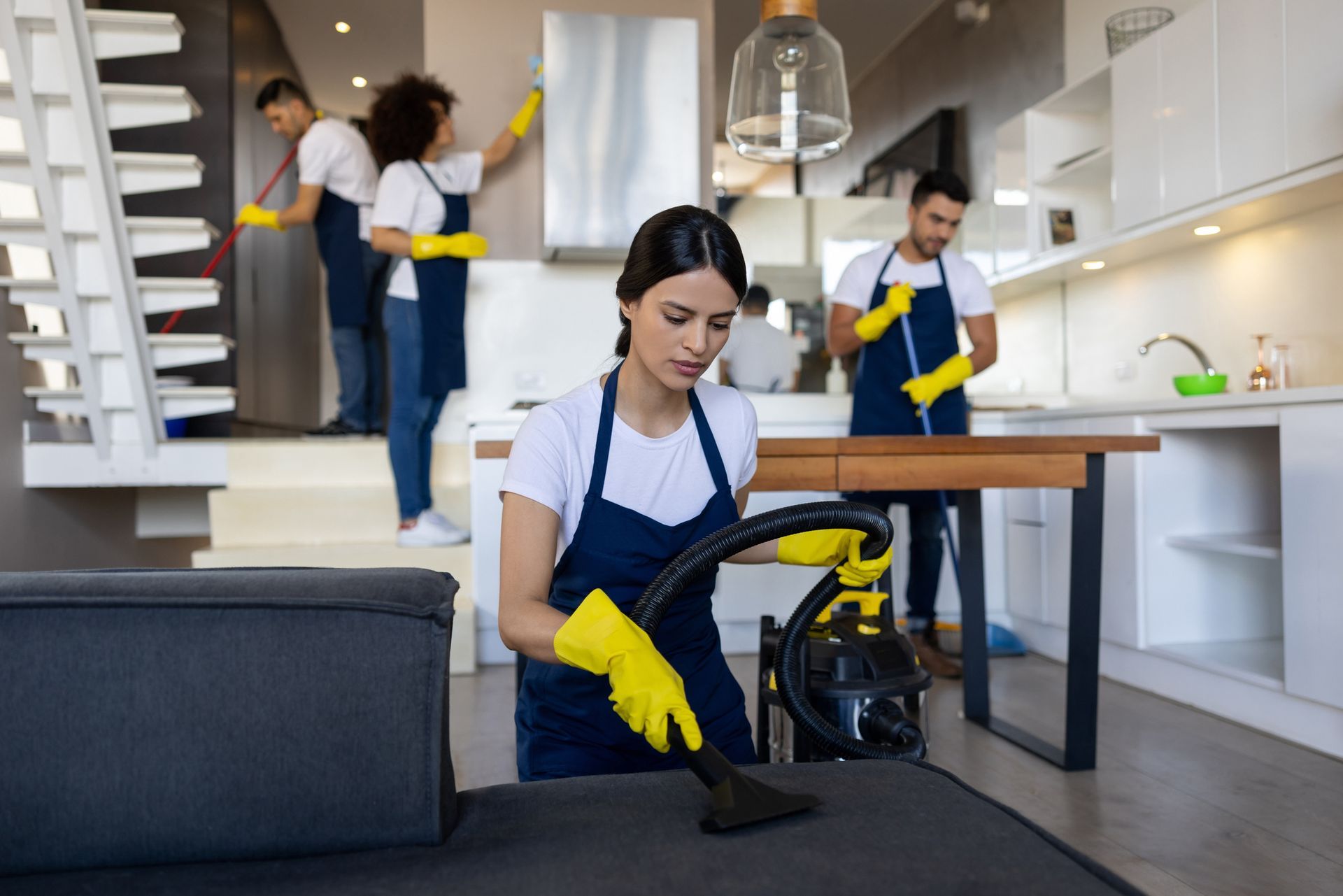 Cleaning crew vacuuming and dusting a living area. One person vacuums a couch, others dust walls and counters.