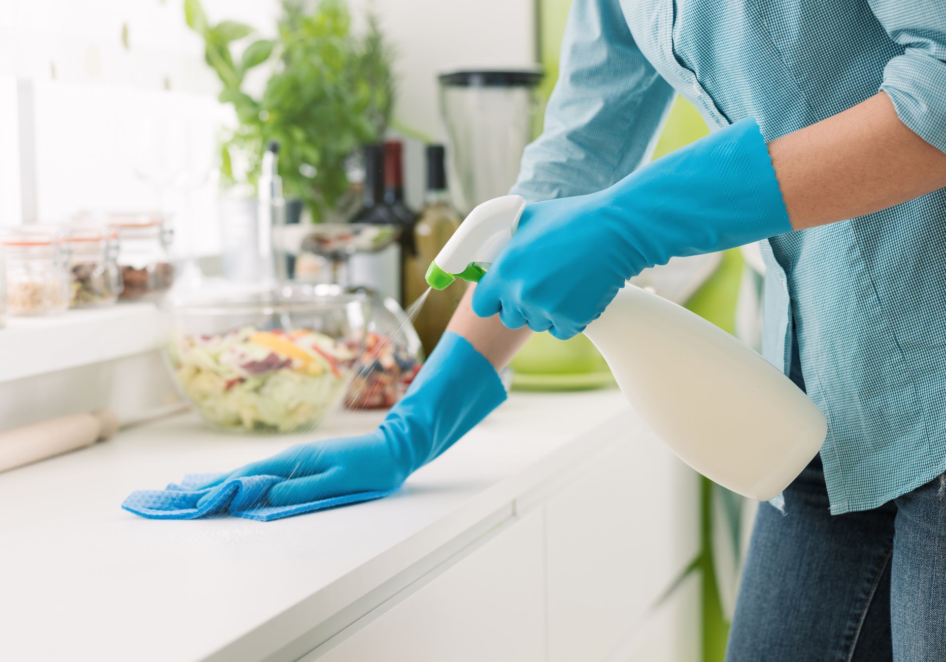 Person wearing blue gloves sprays and wipes a white kitchen countertop.