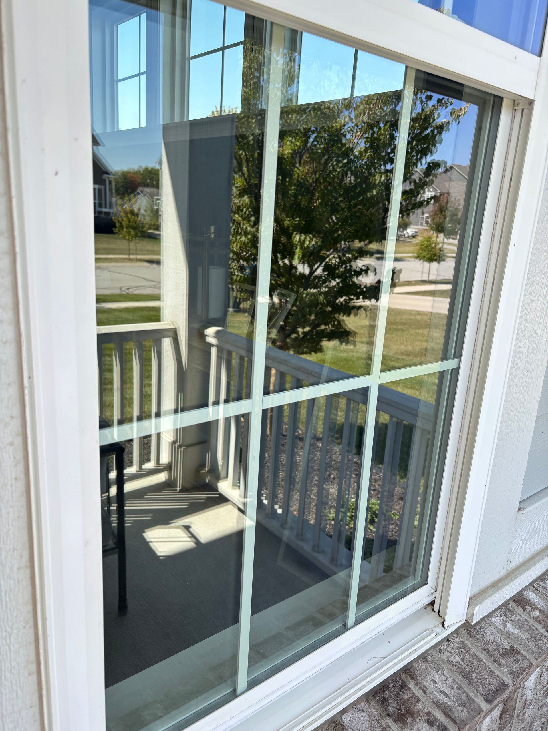 Sliding glass door reflects a porch with a railing and trees in a sunny outdoor setting.