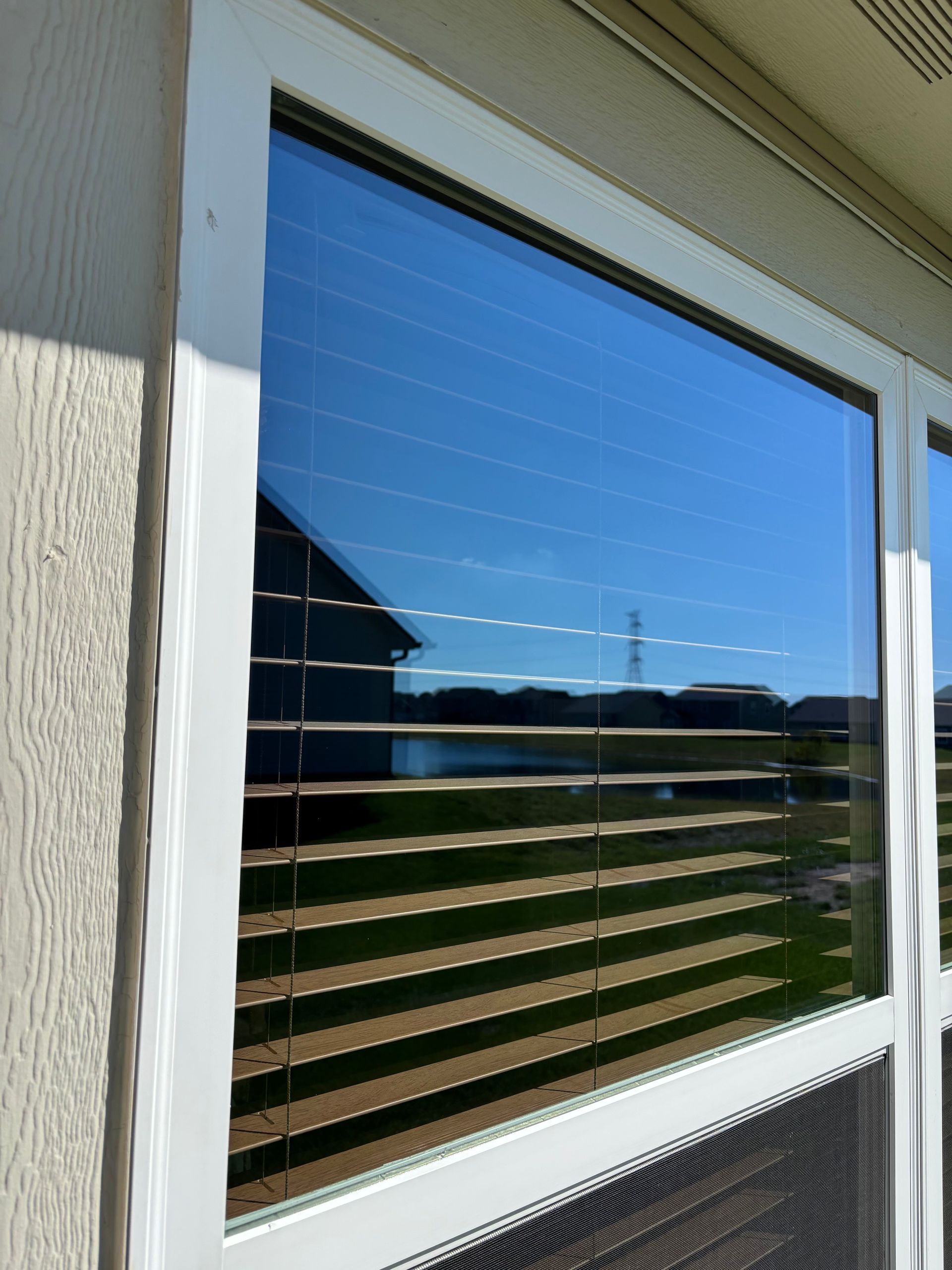 Window with white frame reflecting a blue sky and distant buildings, blinds are closed.