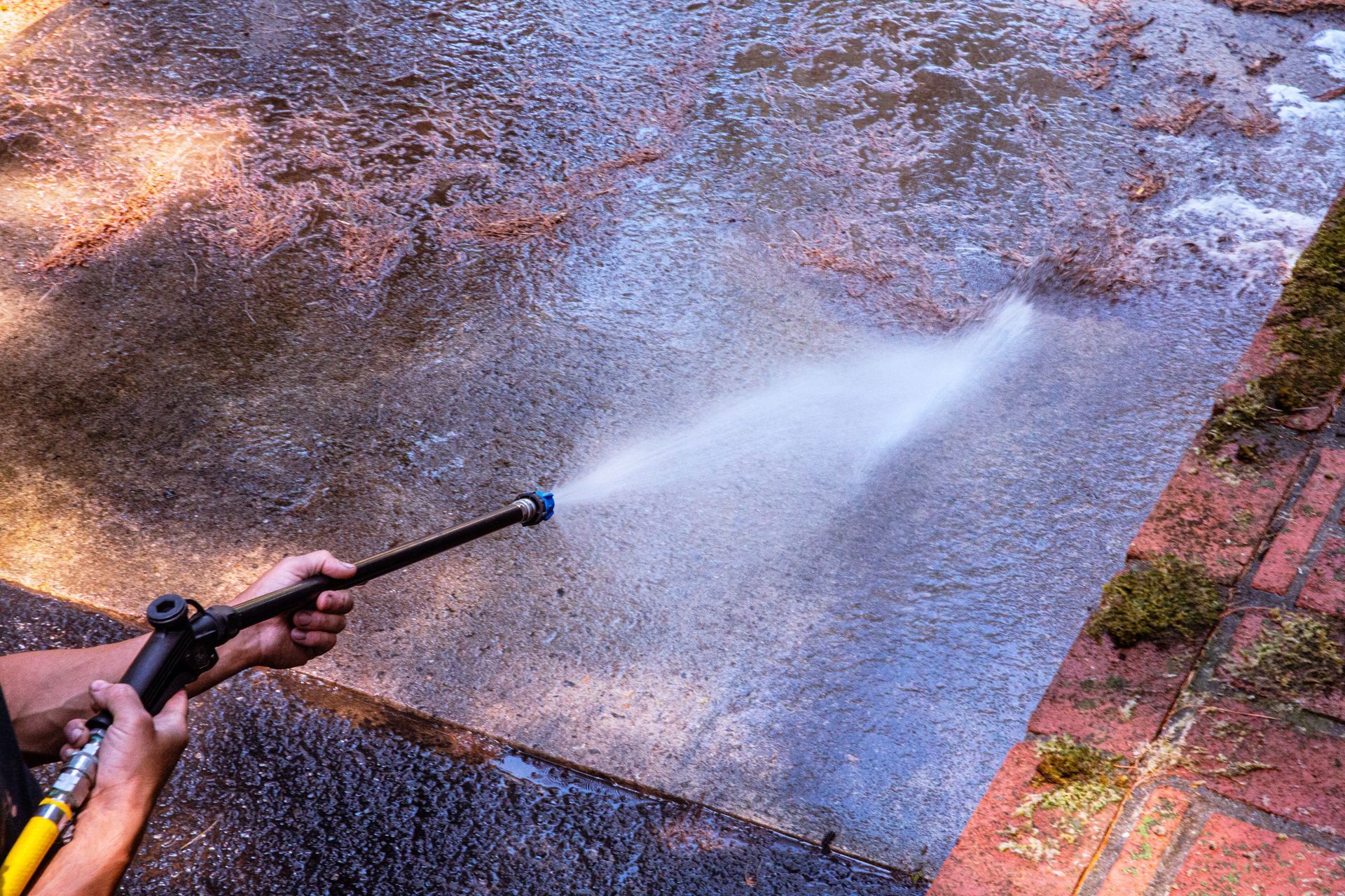 Person pressure washing a concrete surface with a spray of water; near a brick border.