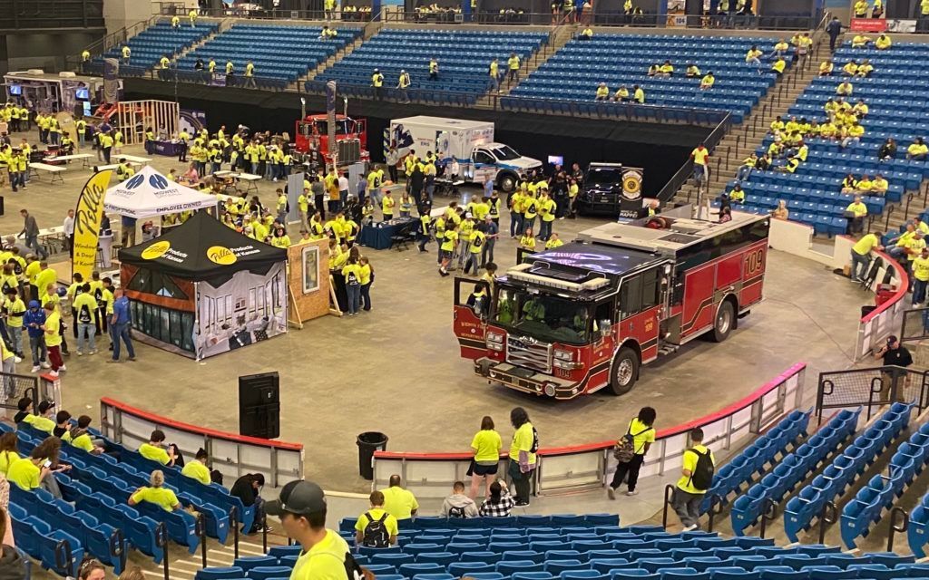 Indoor arena with firefighters and emergency trucks gathered around a central display area in stadium seating