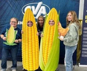 Five people posing with oversized corn cobs in front of a blue event backdrop.