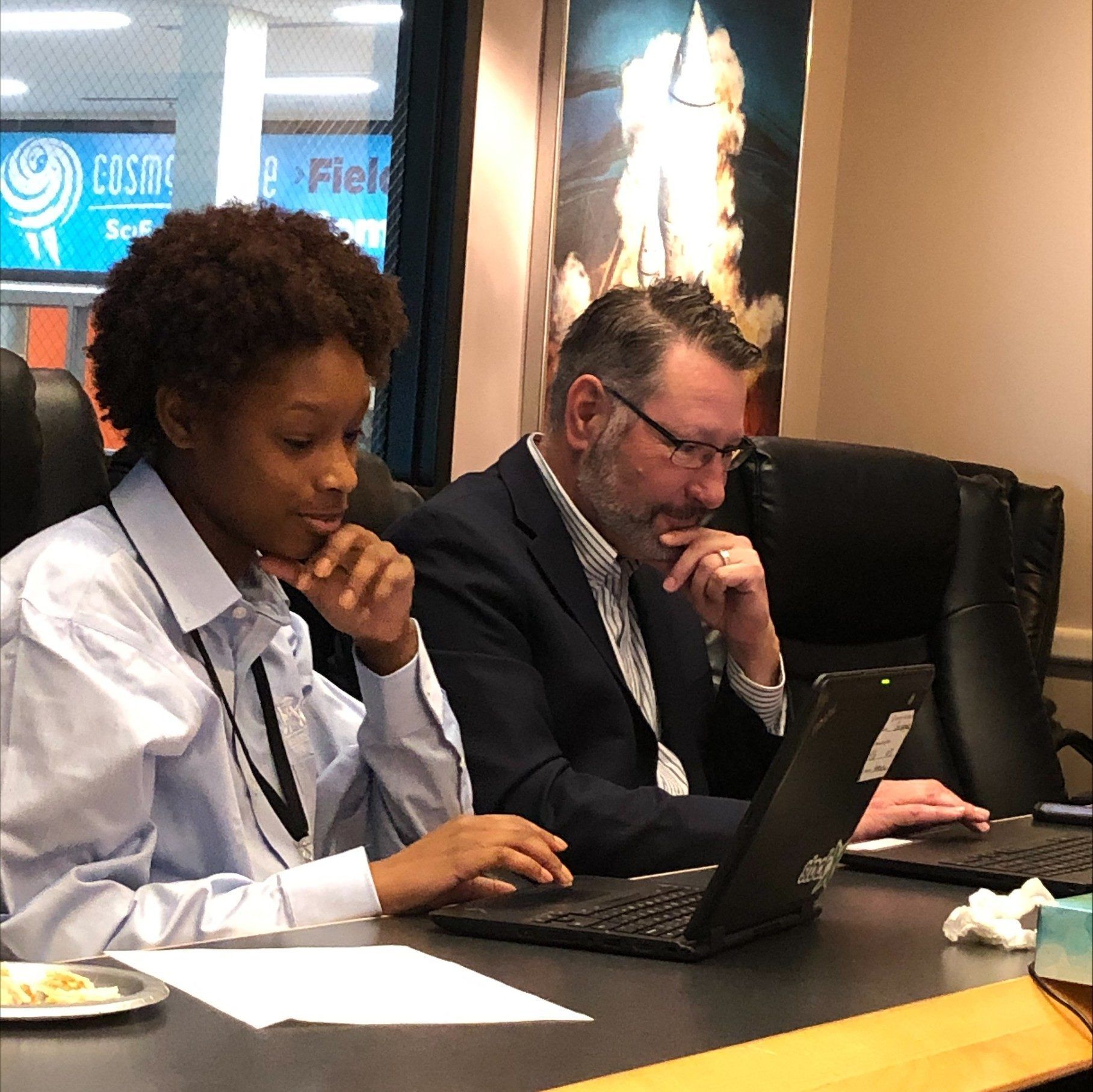 Two people in business attire using laptops at a conference table in a meeting setting