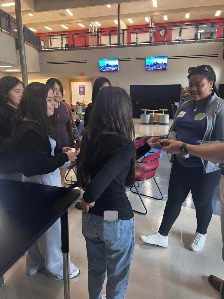 People chatting in a bright lobby, holding colorful items near a counter and two wall-mounted screens.
