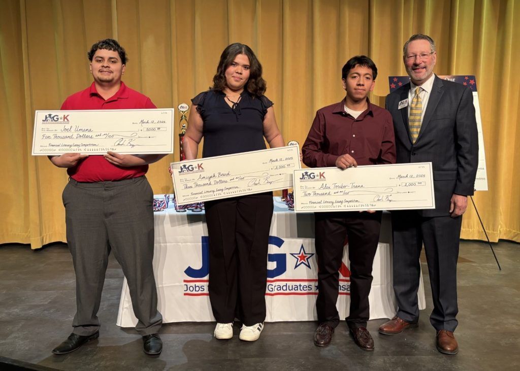 Three people hold oversized checks on a stage with gold curtains; a man in a suit stands at right.