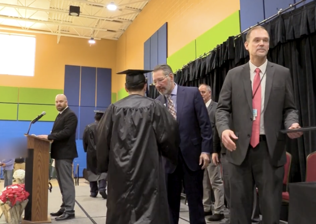 Graduation ceremony with a graduate in cap and gown on stage, officials nearby, and a podium at left