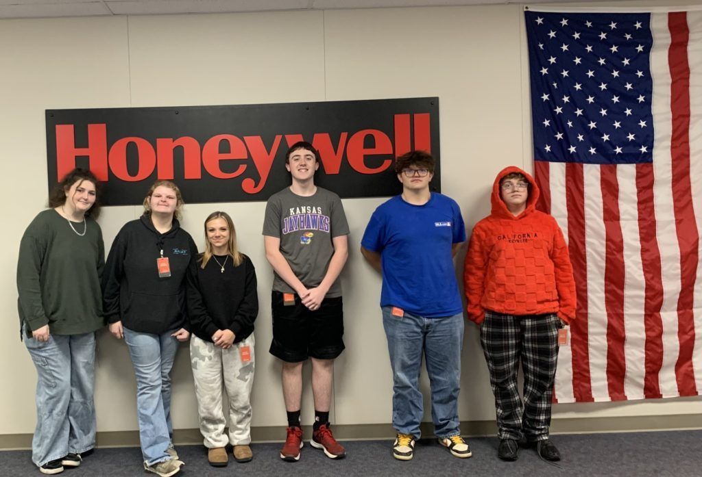 Six people standing indoors in front of a Honeywell sign and a large American flag