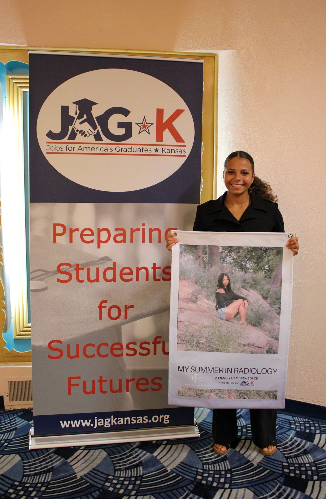 Woman holding a poster beside an NJK banner that reads “Preparing Students for Successful Futures.”