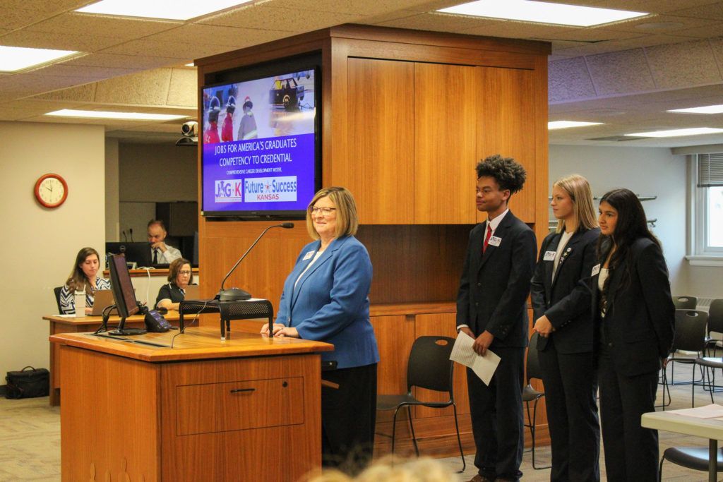 Woman speaking at a podium in a meeting room, with four people standing beside her and a presentation screen behind.