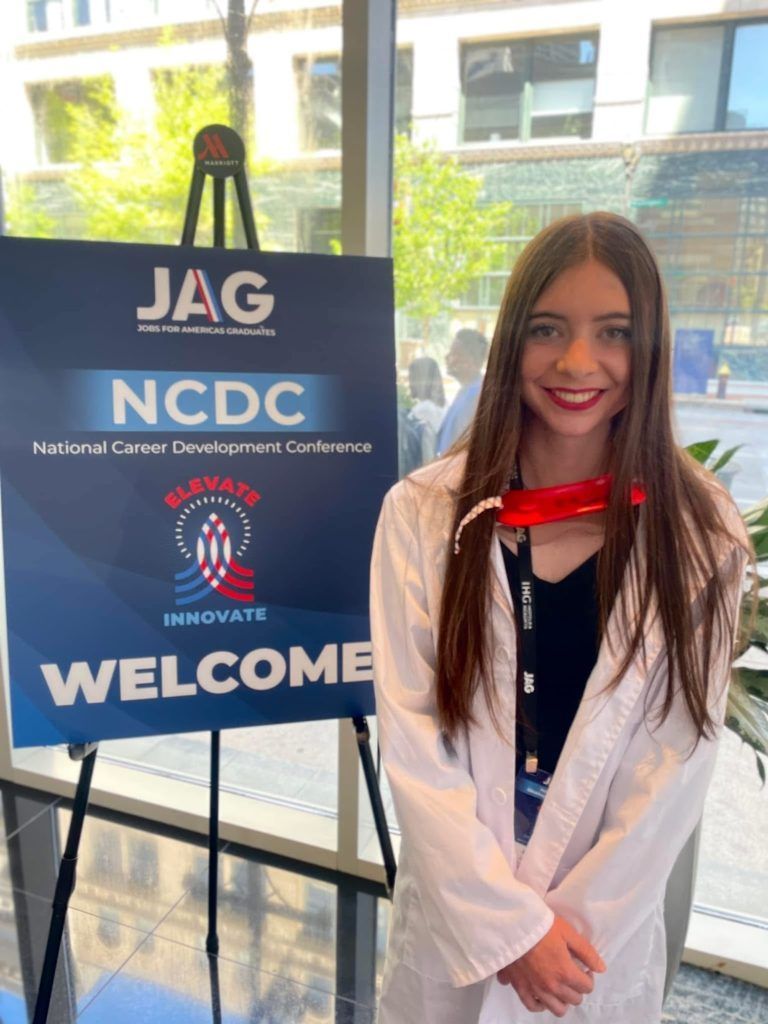 Woman in white coat beside JAG NCDC welcome sign at an indoor conference booth.