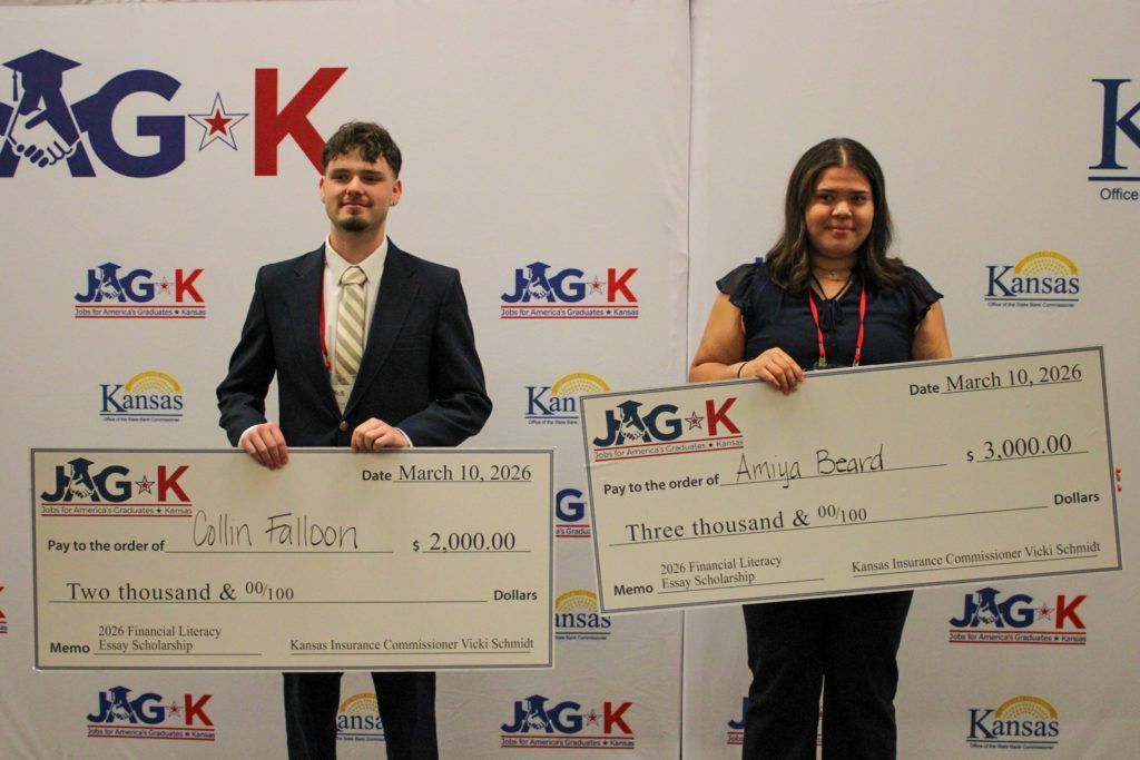 Two people hold oversized checks at an awards event with Kansas and JG+K logos behind them.