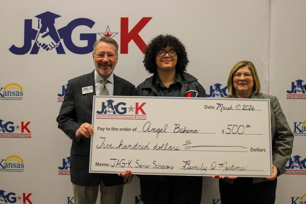 Three people hold a large ceremonial check in front of a JAG-K backdrop.