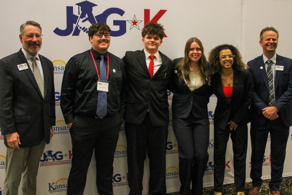 Six people in suits pose in front of a JAG-K banner at an indoor event