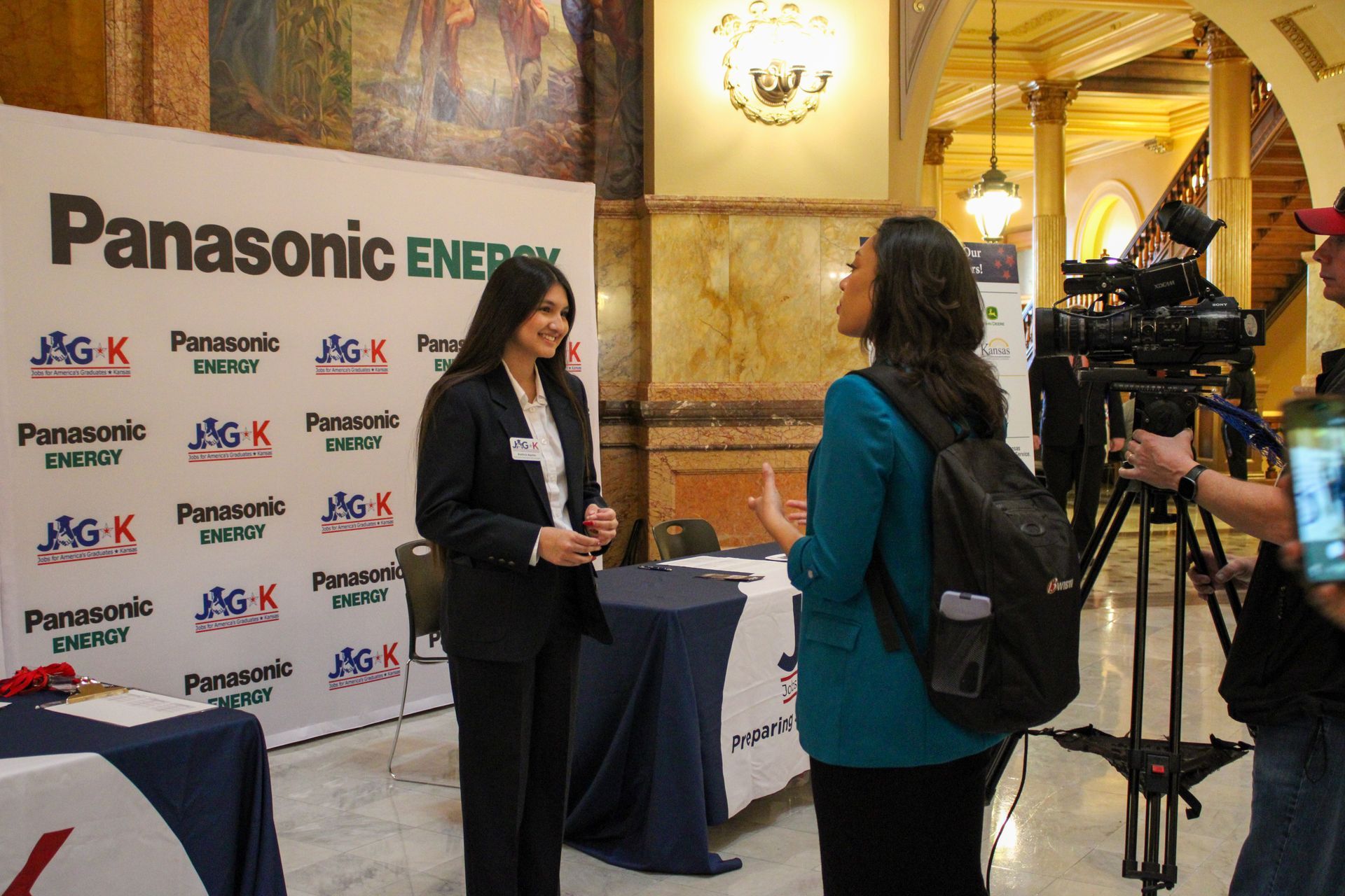 Two women speaking at a Panasonic Energy event booth with logos and a table display