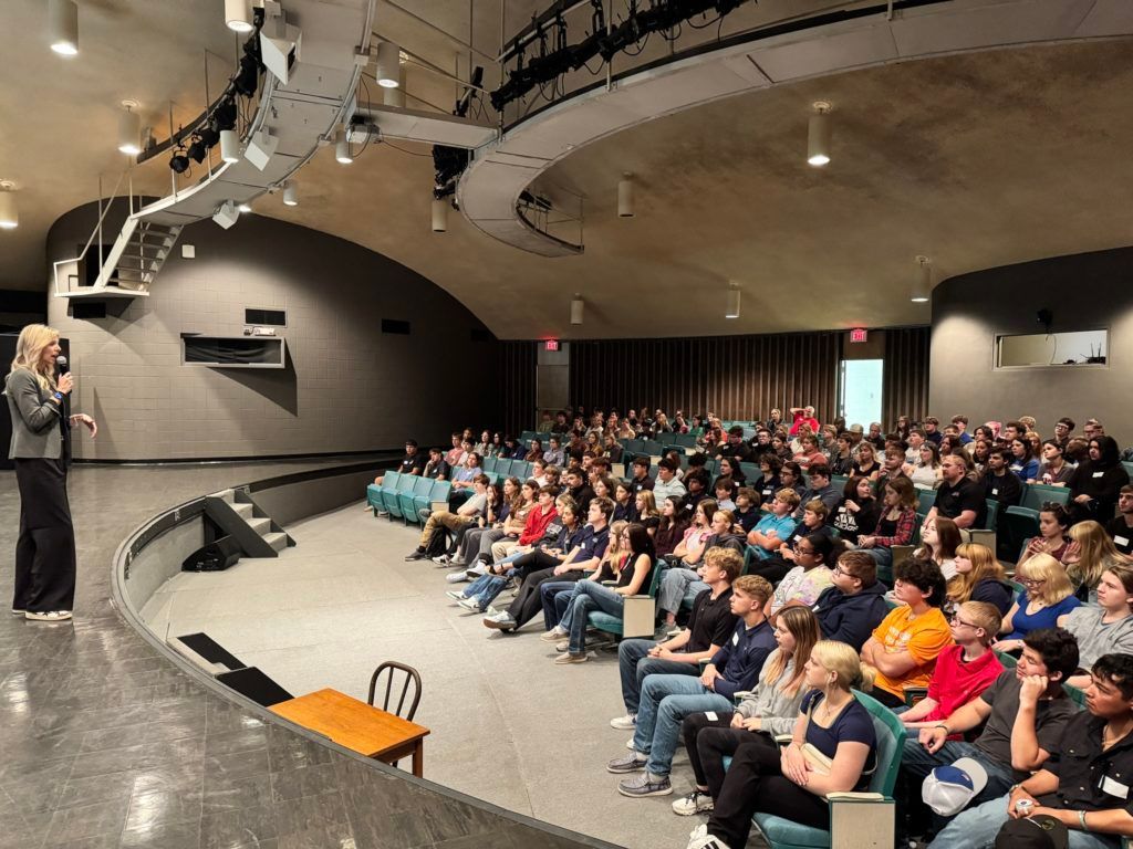 Speaker addressing seated audience in a large auditorium with curved ceiling and stage lighting