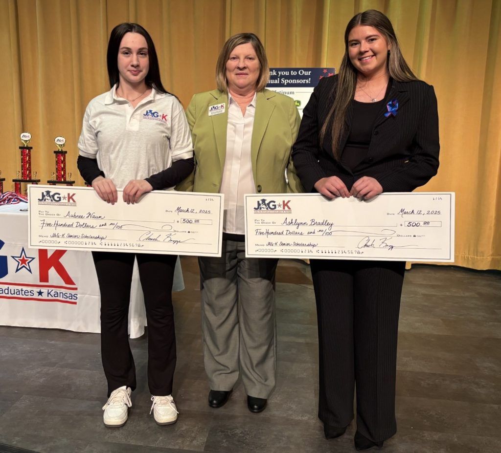 Three people holding oversized checks at an awards ceremony, standing indoors beside trophies.
