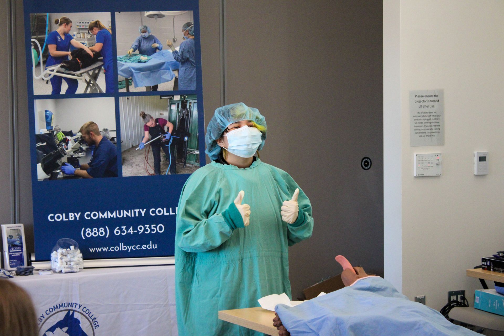 Doctor in green scrubs and mask giving a thumbs-up beside a patient in a clinic exam room.