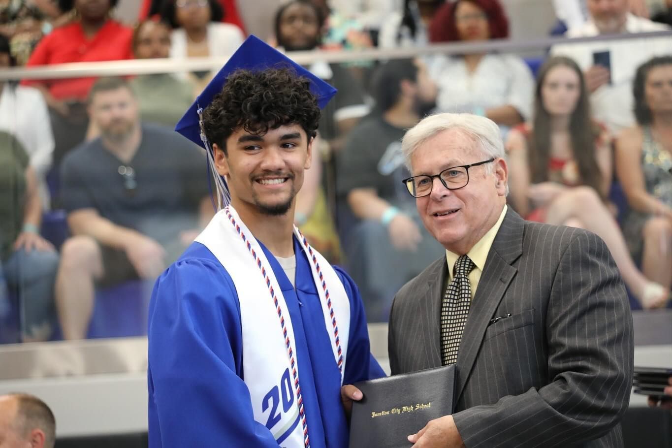 Graduation ceremony: student in blue cap and gown holds diploma with an older man in a suit on stage