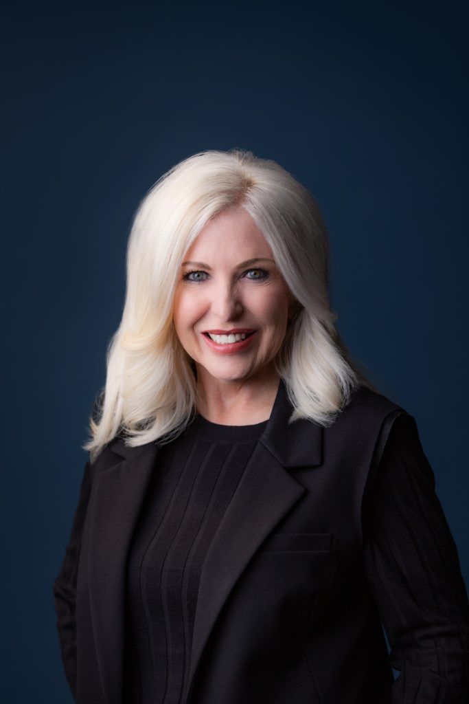 Smiling woman with blonde hair wearing a black blazer against a dark blue background