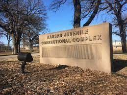 Kansas Juvenile Correctional Complex sign in a leaf-covered outdoor setting with bare trees and blue sky