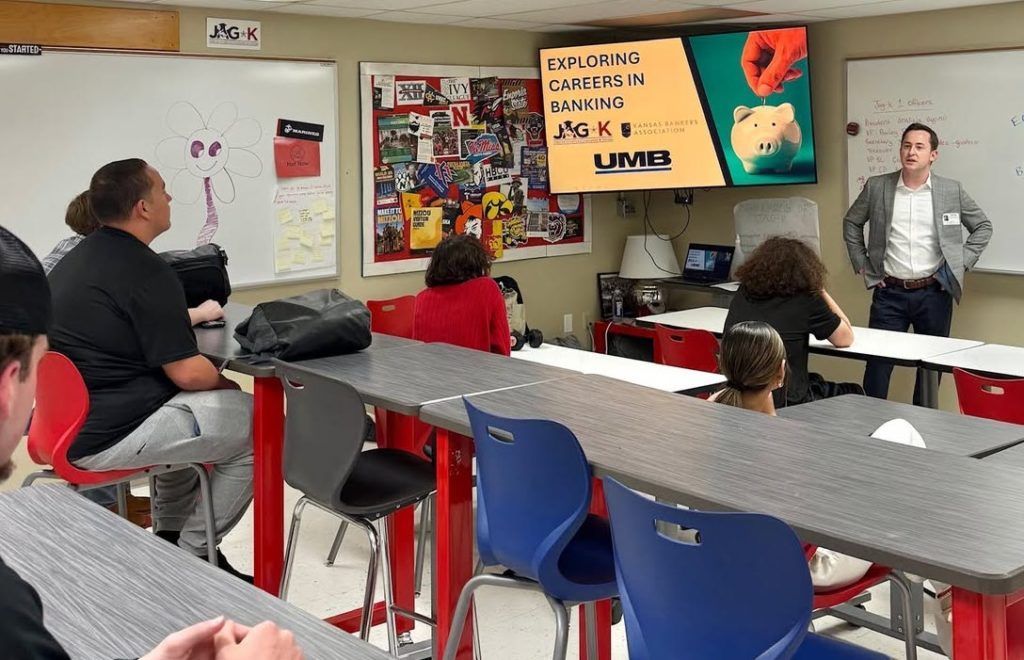 Classroom with students seated at tables while a teacher presents beside a display screen
