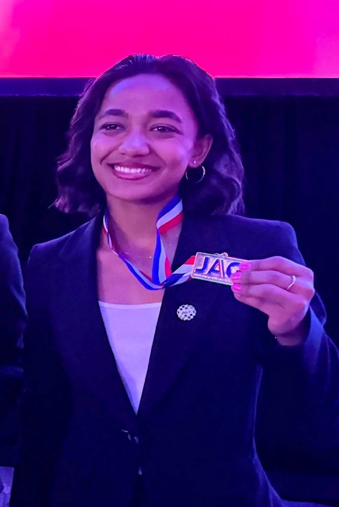 Smiling person in a black blazer holds up a medal with a red-white-blue ribbon