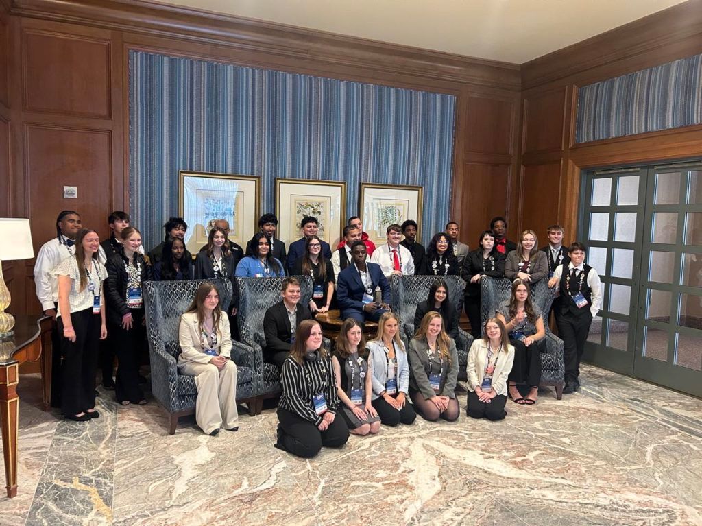Large group posing in a wood-paneled room with framed artwork and blue-striped curtains
