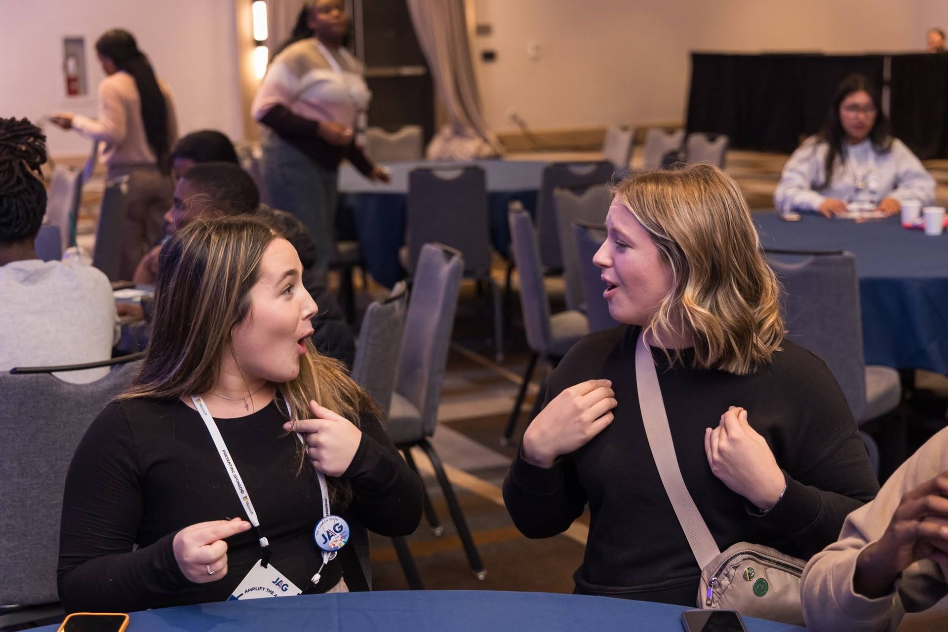 Two women chatting at a conference table in a meeting room, with laptops and people in the background.