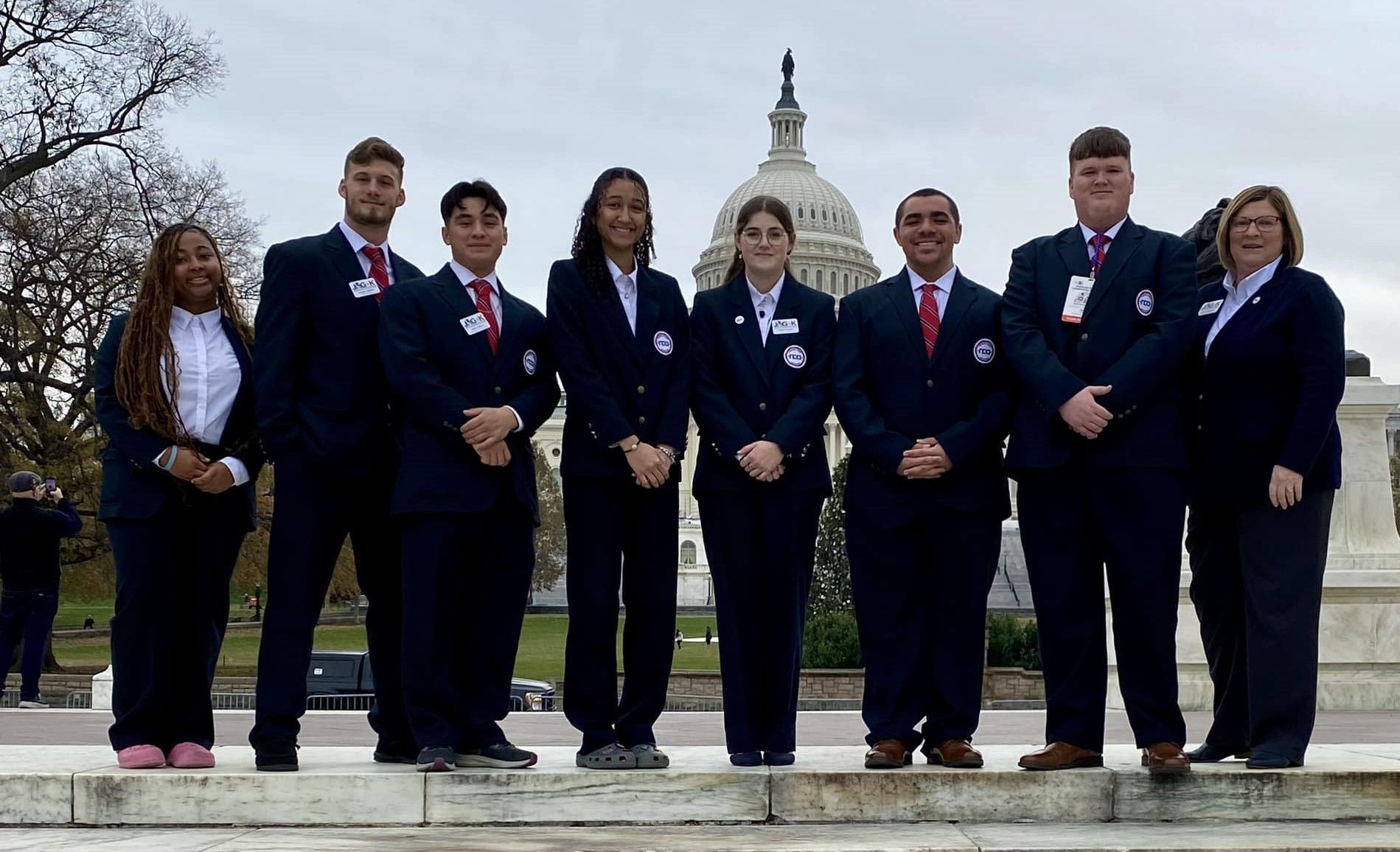 Nine people in suits pose outdoors in front of a capitol building, holding certificates and medals.
