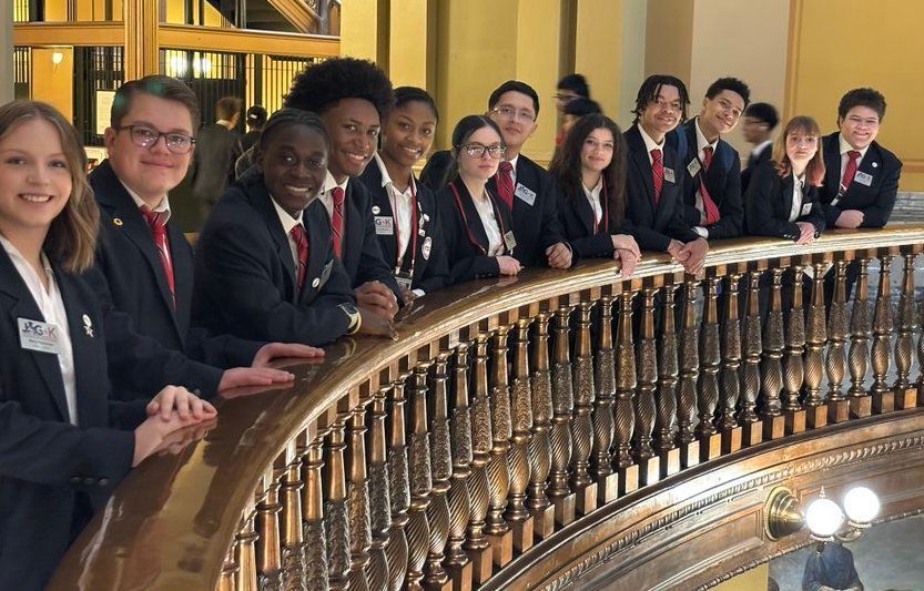 Group of students in dark blazers and red ties standing along an ornate balcony railing indoors
