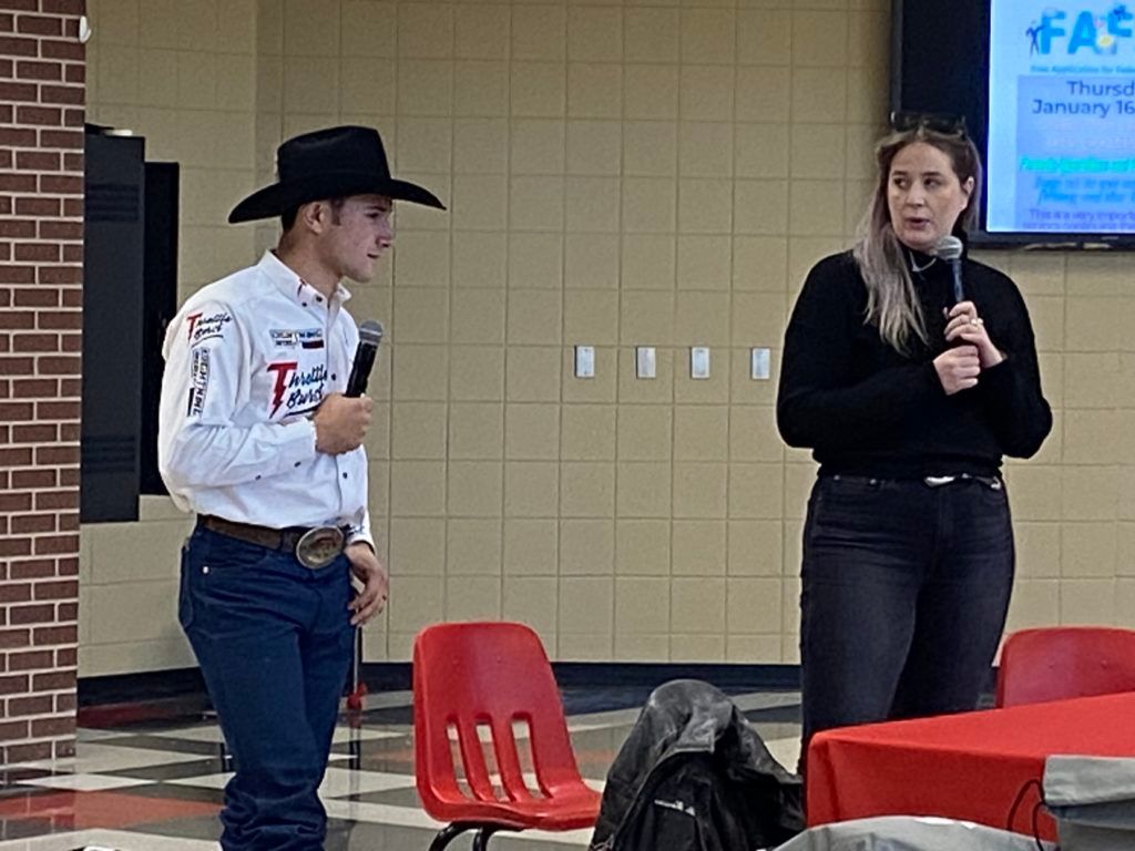 Two people speaking indoors near red chairs, one holding a microphone and wearing a black cowboy hat