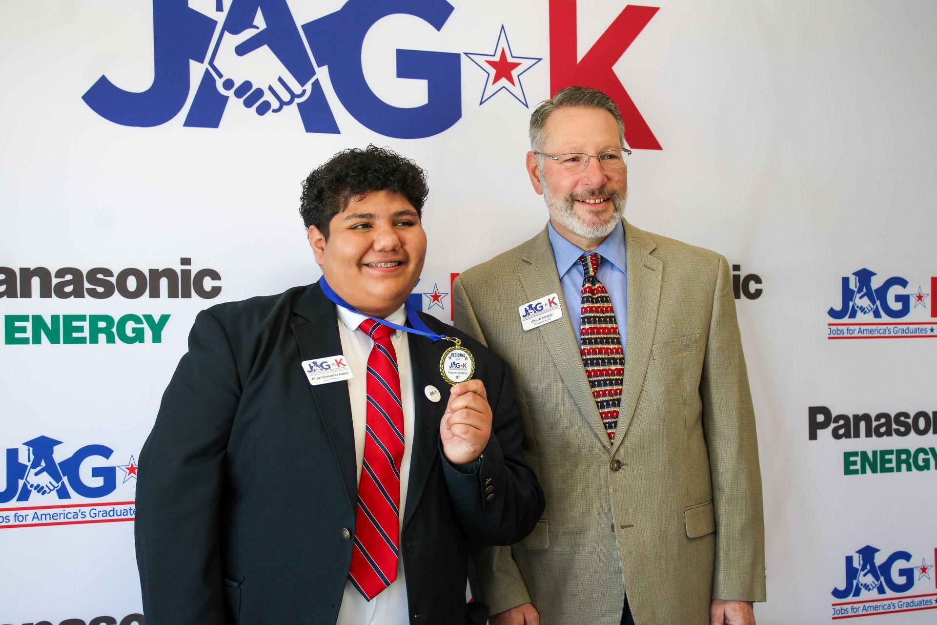 Two men in suits posing at a JAGX event backdrop, one holding an award and giving a thumbs-up.