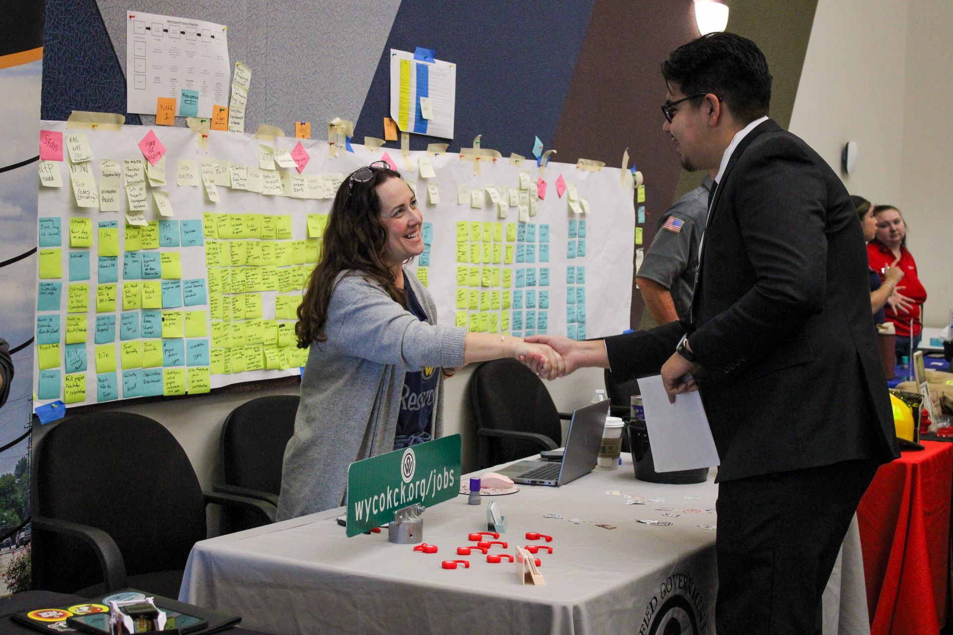 Two people shake hands at a booth with colorful sticky notes and a table sign.