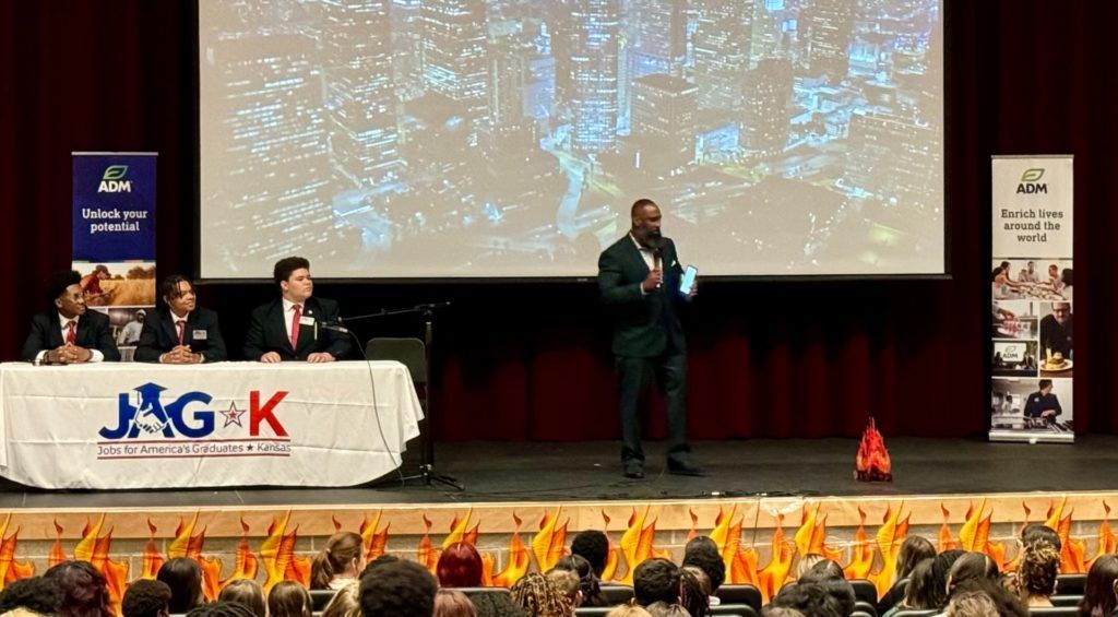 Conference stage with speakers at a podium, large screen backdrop, and audience holding up orange lights