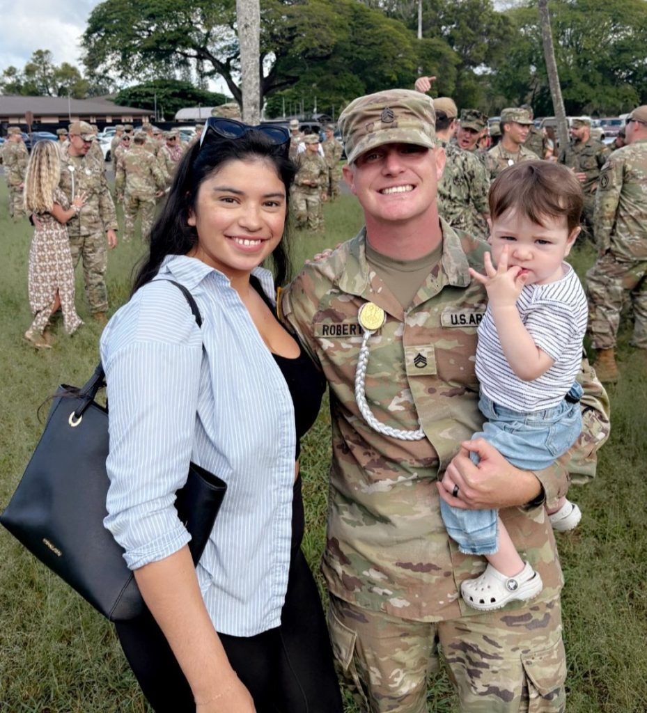 Smiling soldier in camouflage holding a child, standing with a woman outdoors at a military gathering.