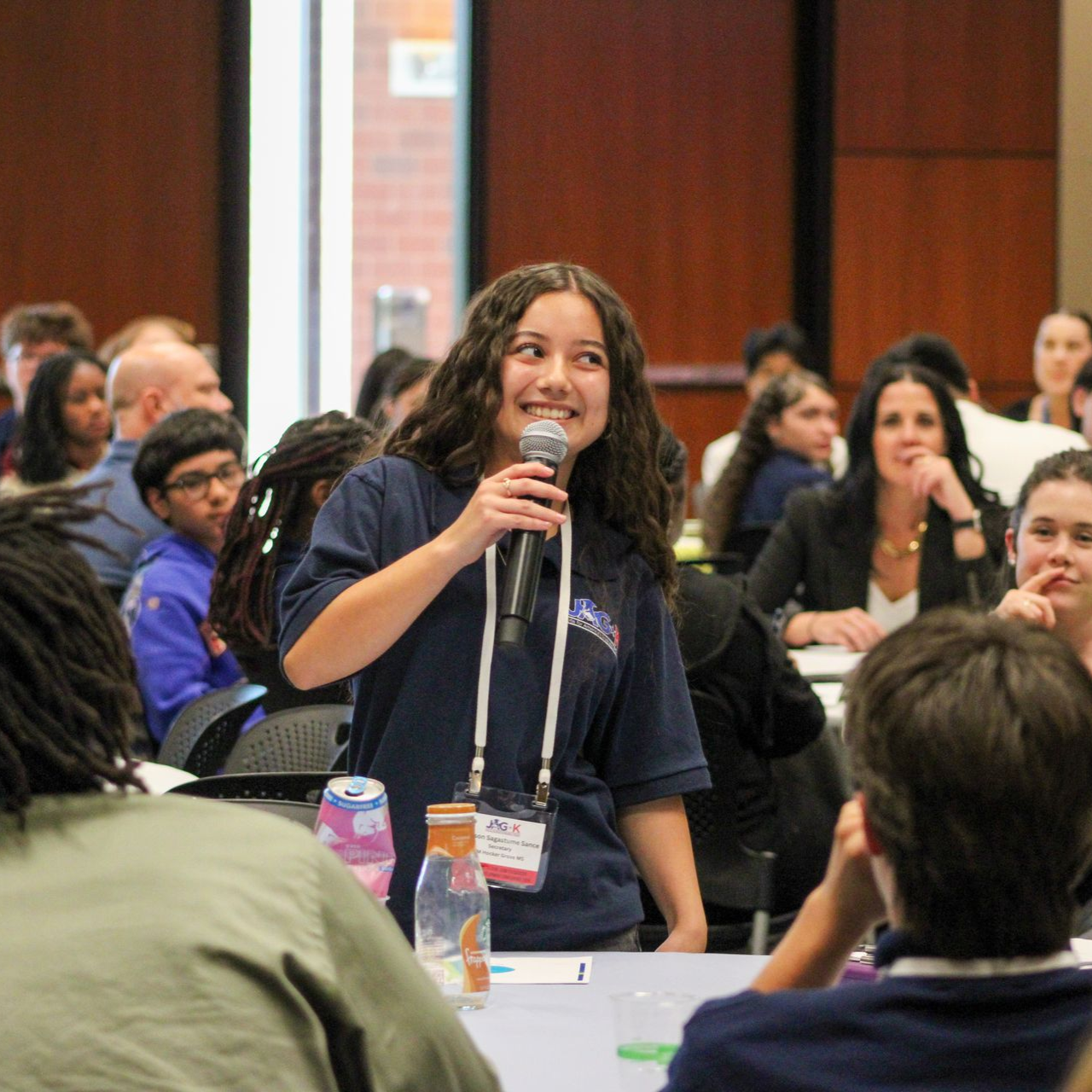 Person speaking into a microphone at a conference, with seated audience members in the background.