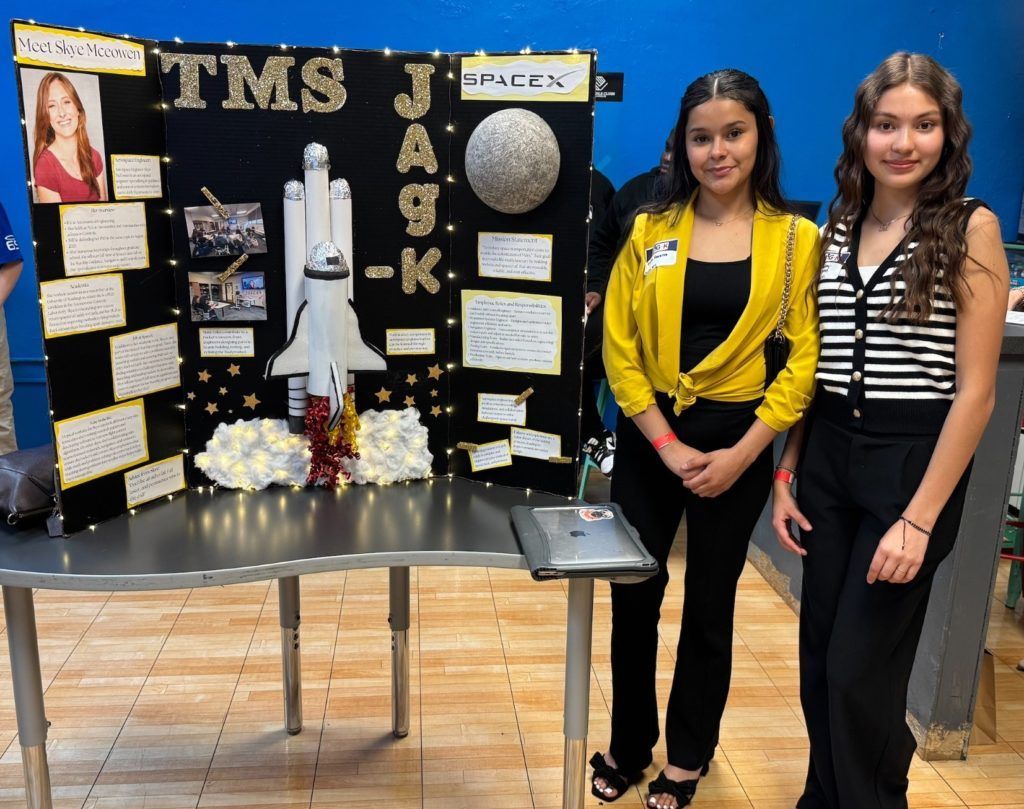 Two girls stand beside a science fair display board featuring a rocket and space-themed decorations.
