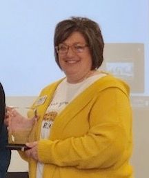 Woman in a yellow sweater smiling while holding a plaque at an indoor ceremony.
