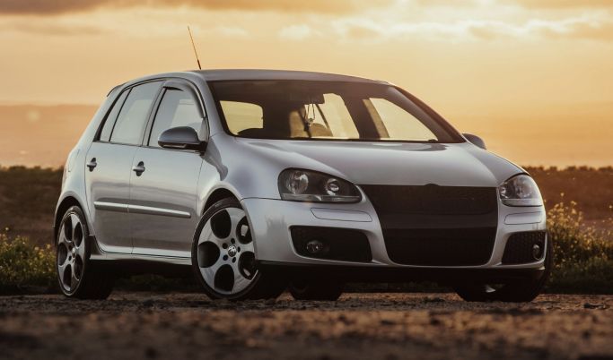 A silver car is parked on a dirt road at sunset.