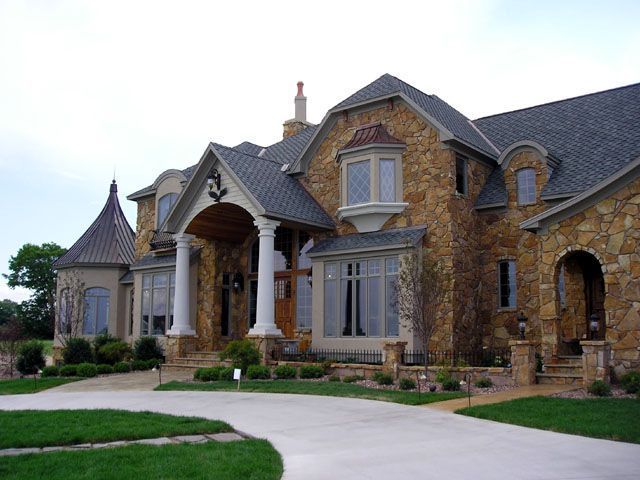 Stone-clad mansion with columns, arched doorway, and driveway; overcast sky.