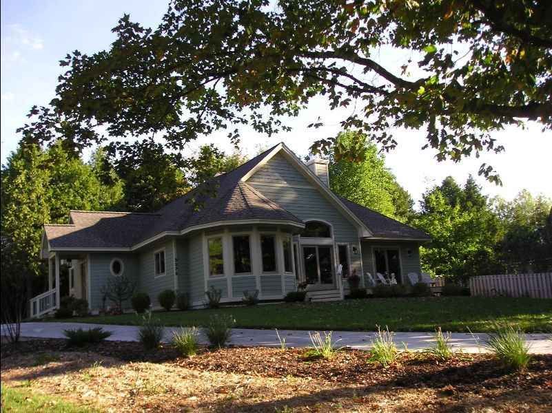 Light blue house with curved window and porch under a tree. Green lawn, blue sky.