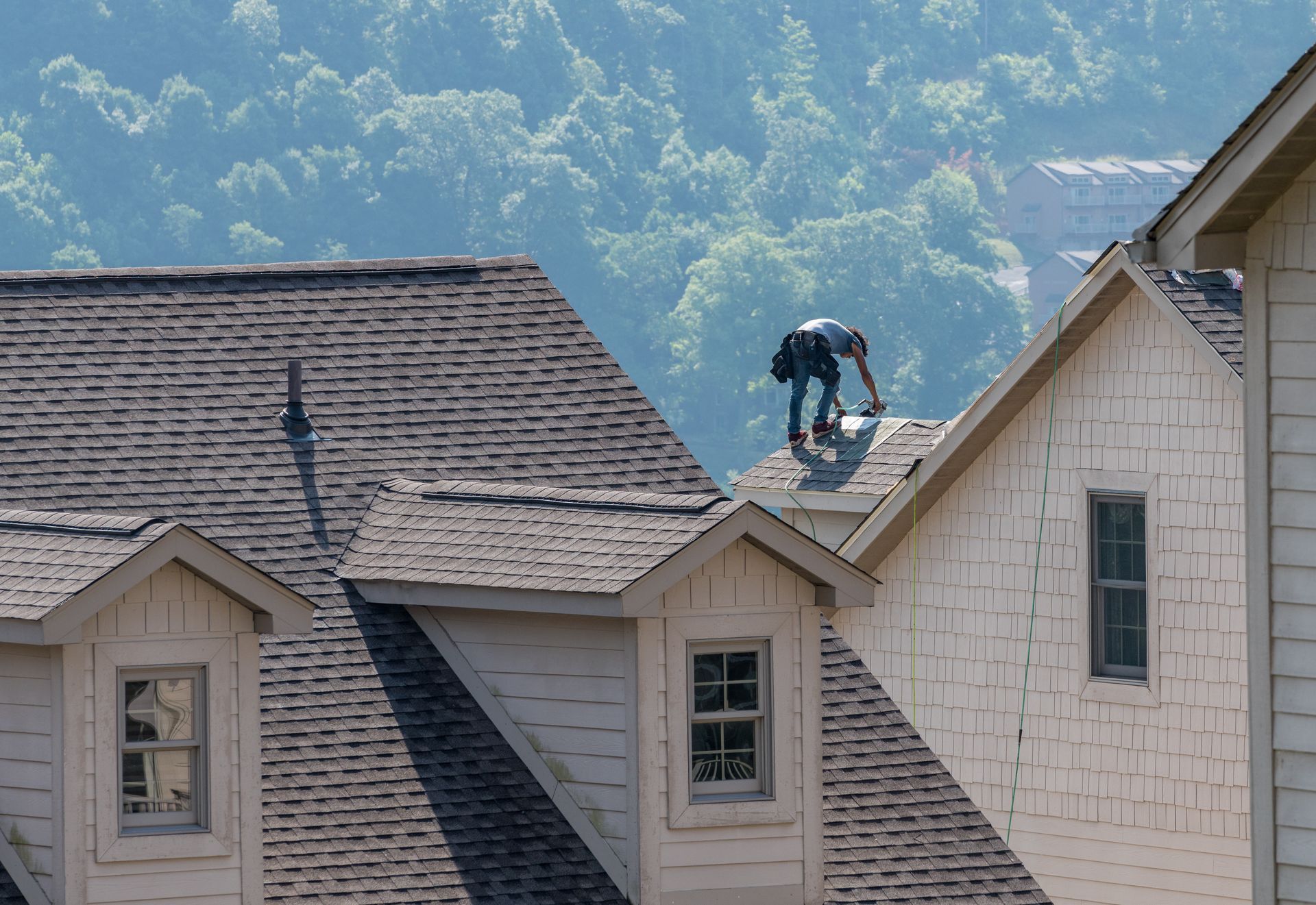 Worker on a rooftop shingling a house, with wooded hills in the background.