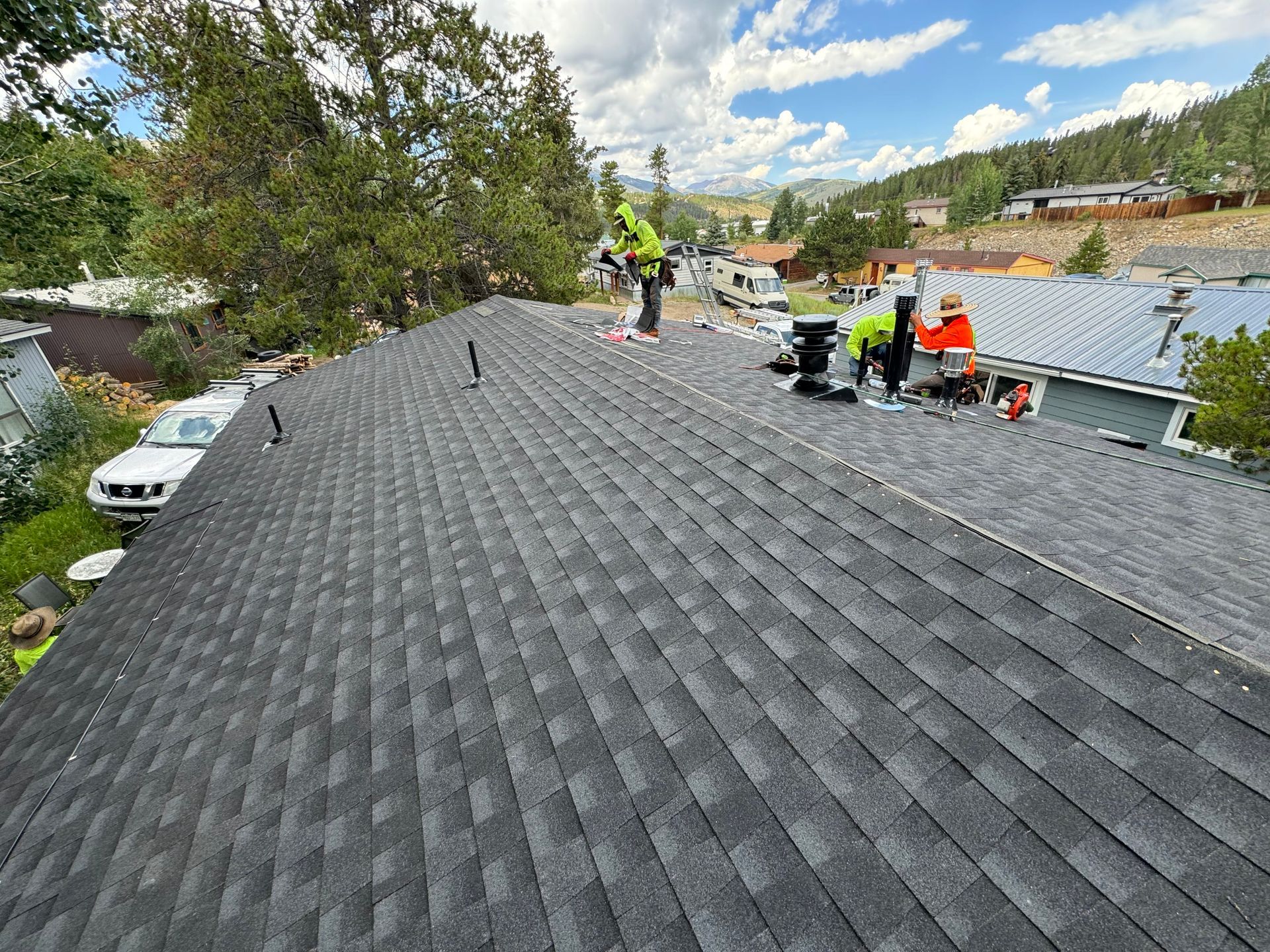 Workers on a gray shingled roof, with tools and materials, overlooking a neighborhood under a cloudy sky Workers on a gray shingled roof, with tools and materials, overlooking a neighborhood under a cloudy sky