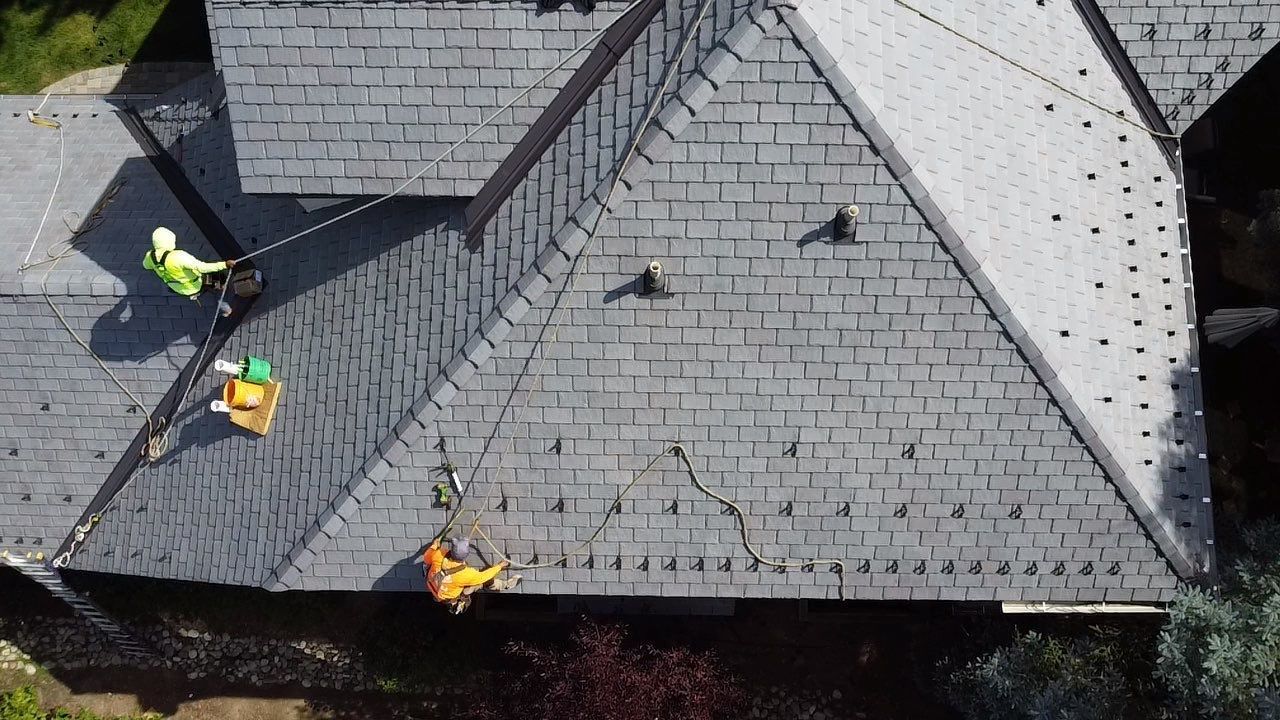 Roofers working on a gray shingled roof with several tools and safety vests visible