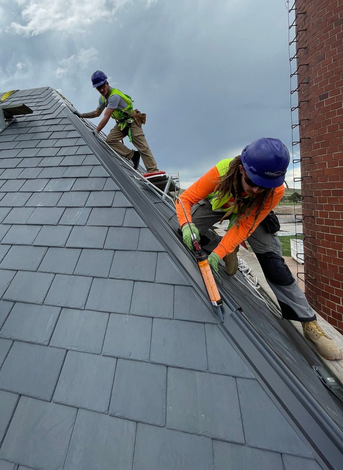 Workers in helmets and safety vests repair a steep slate roof with harnesses.