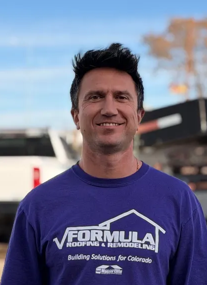 Man in purple shirt smiling in front of truck and building with blue sky.