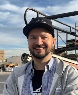 Man in gray hoodie and baseball cap smiles outdoors, arms crossed, with building and cloudy sky in background.