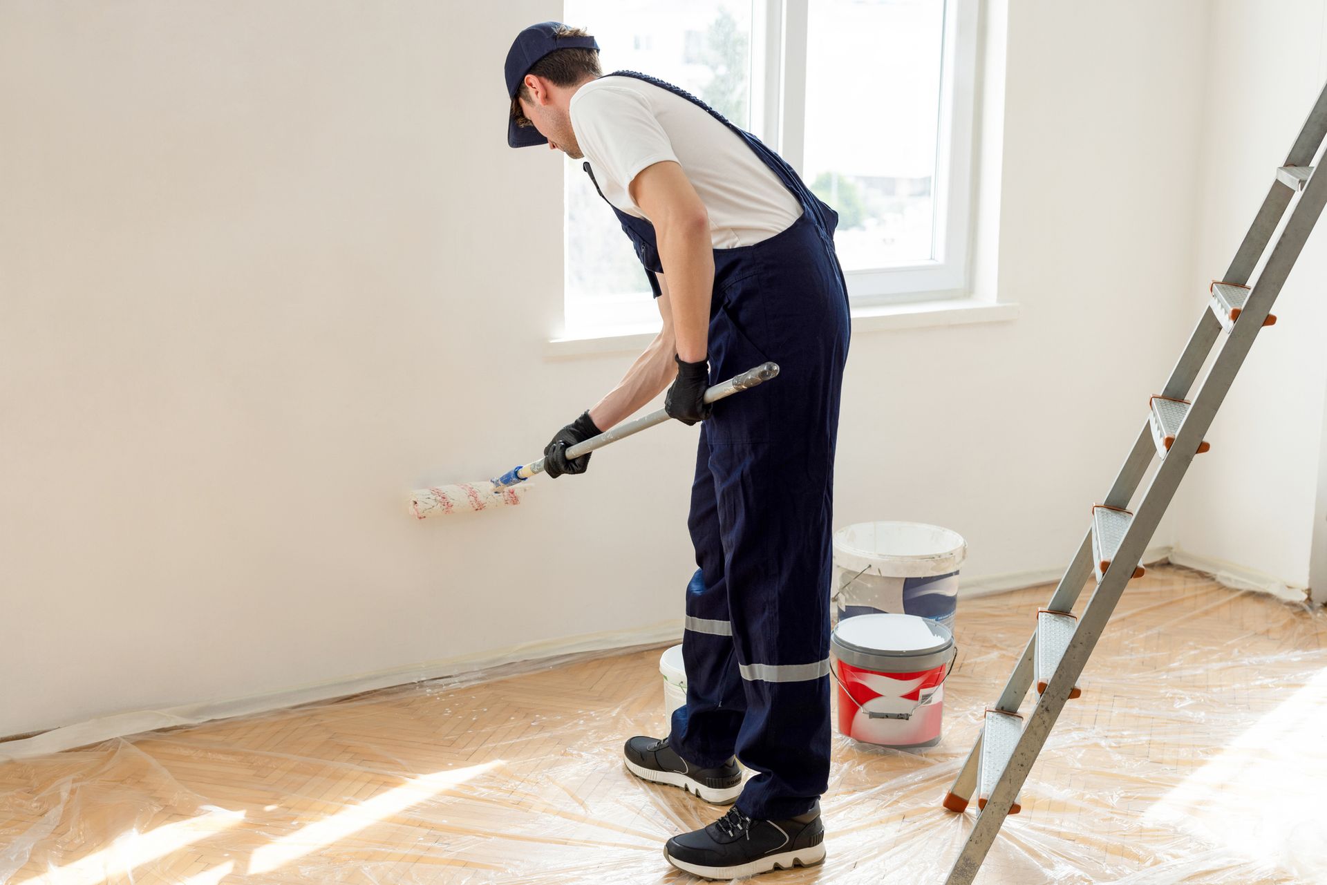 A young male painting contractor wearing a hat and coveralls is painting a room.