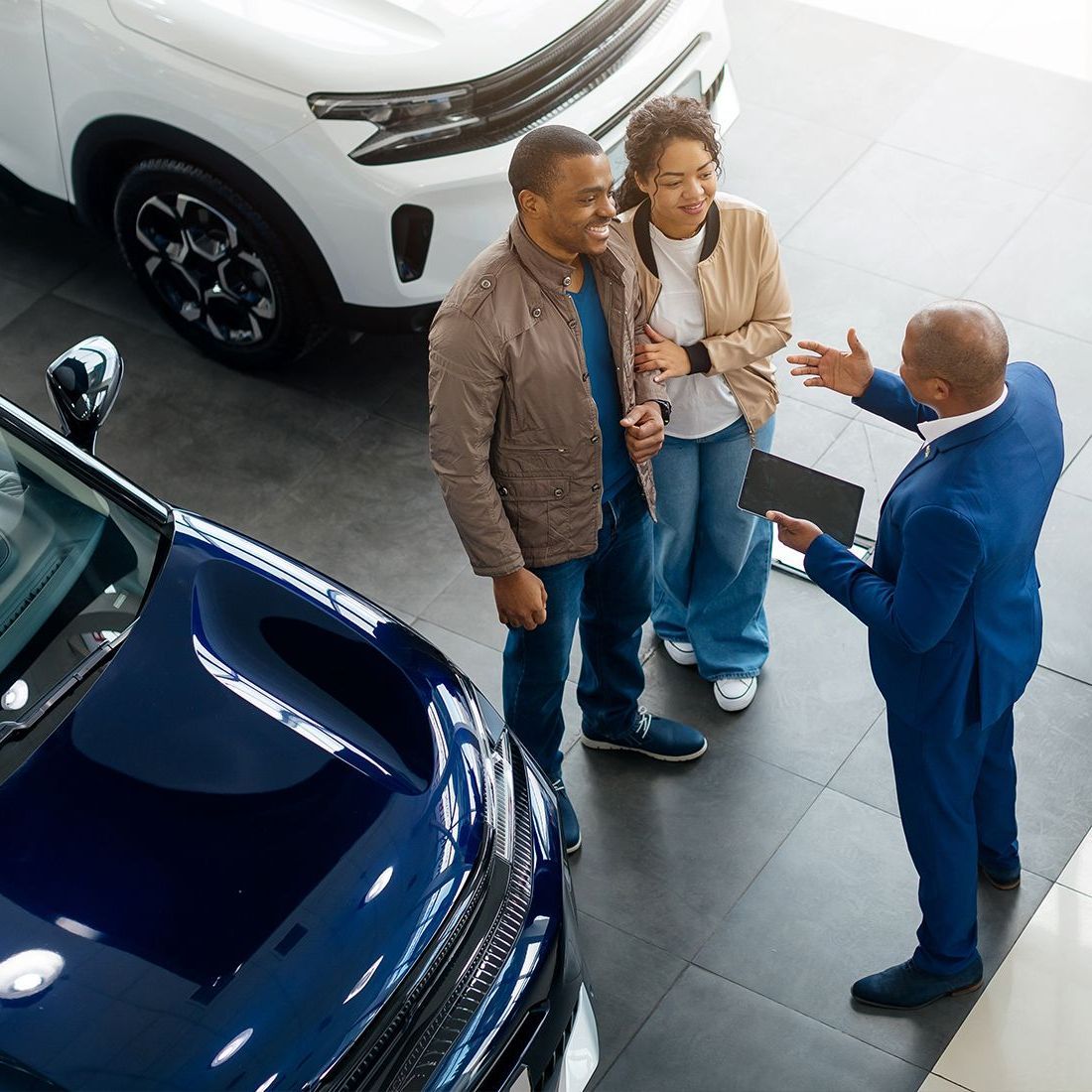 A car salesperson in a blue suit gestures towards a couple in a car showroom. They are looking at cars, smiling.